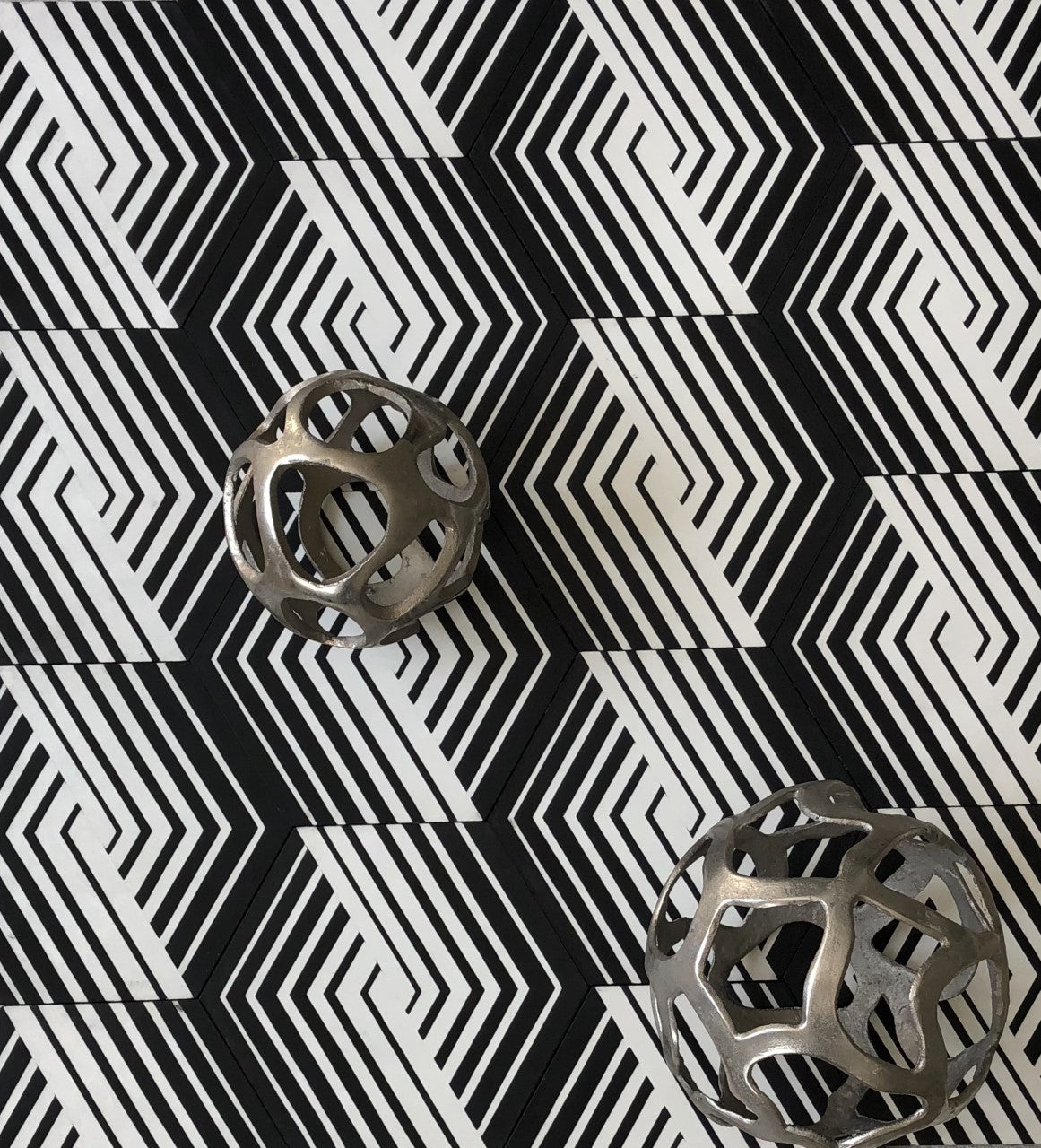 Two metallic, hollow spheres rest on a black-and-white geometric, zigzag patterned surface, creating an optical illusion effect.