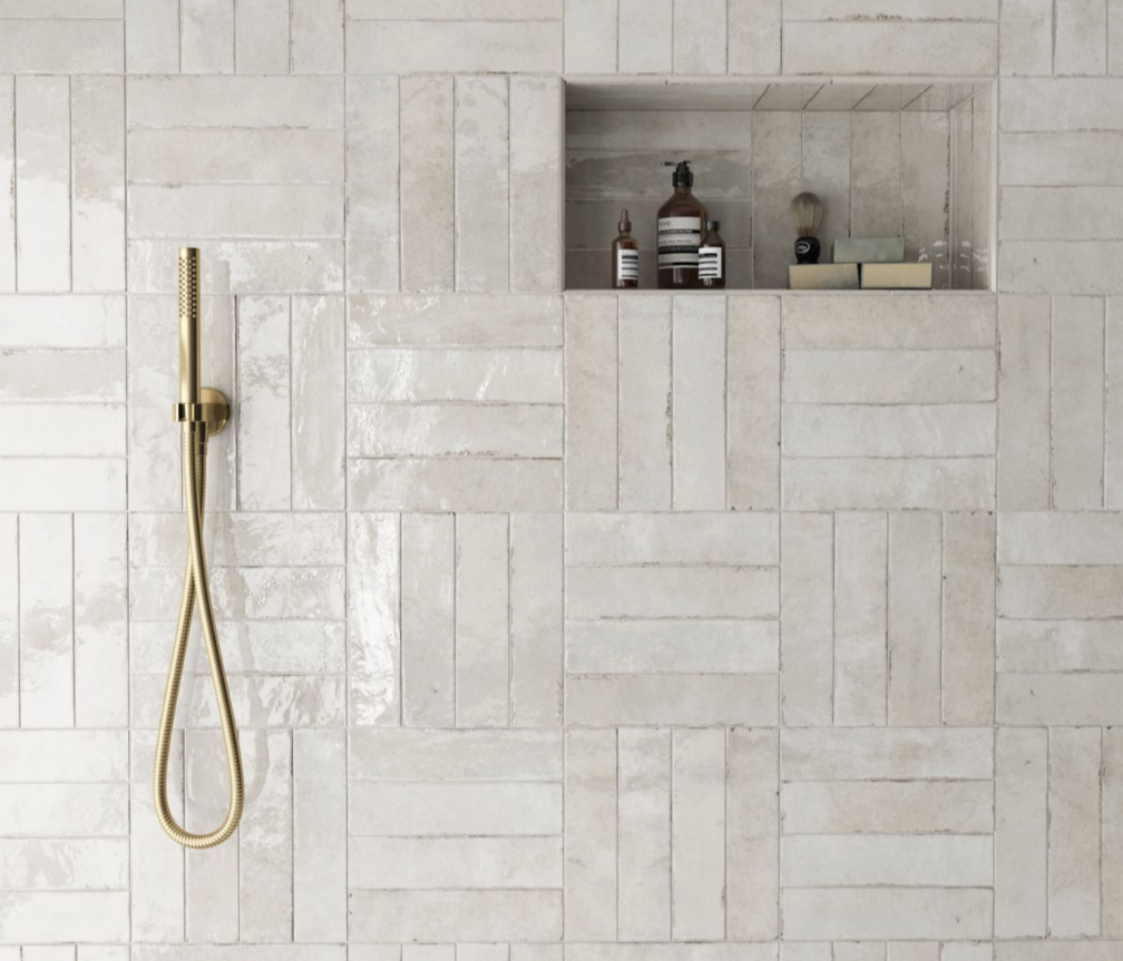 Close-up of a shower wall with glossy beige tiles in a geometric pattern, a brass handheld shower, and a built-in shelf holding toiletries like bottles, a shaving brush, and soap.