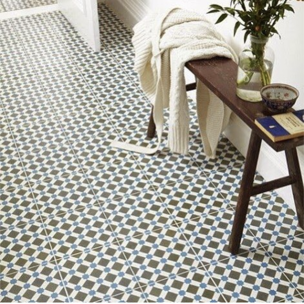 A geometric-patterned tile floor with a wooden bench holding a white knit sweater, a potted plant, a bowl, and a closed notebook placed against a white wall.