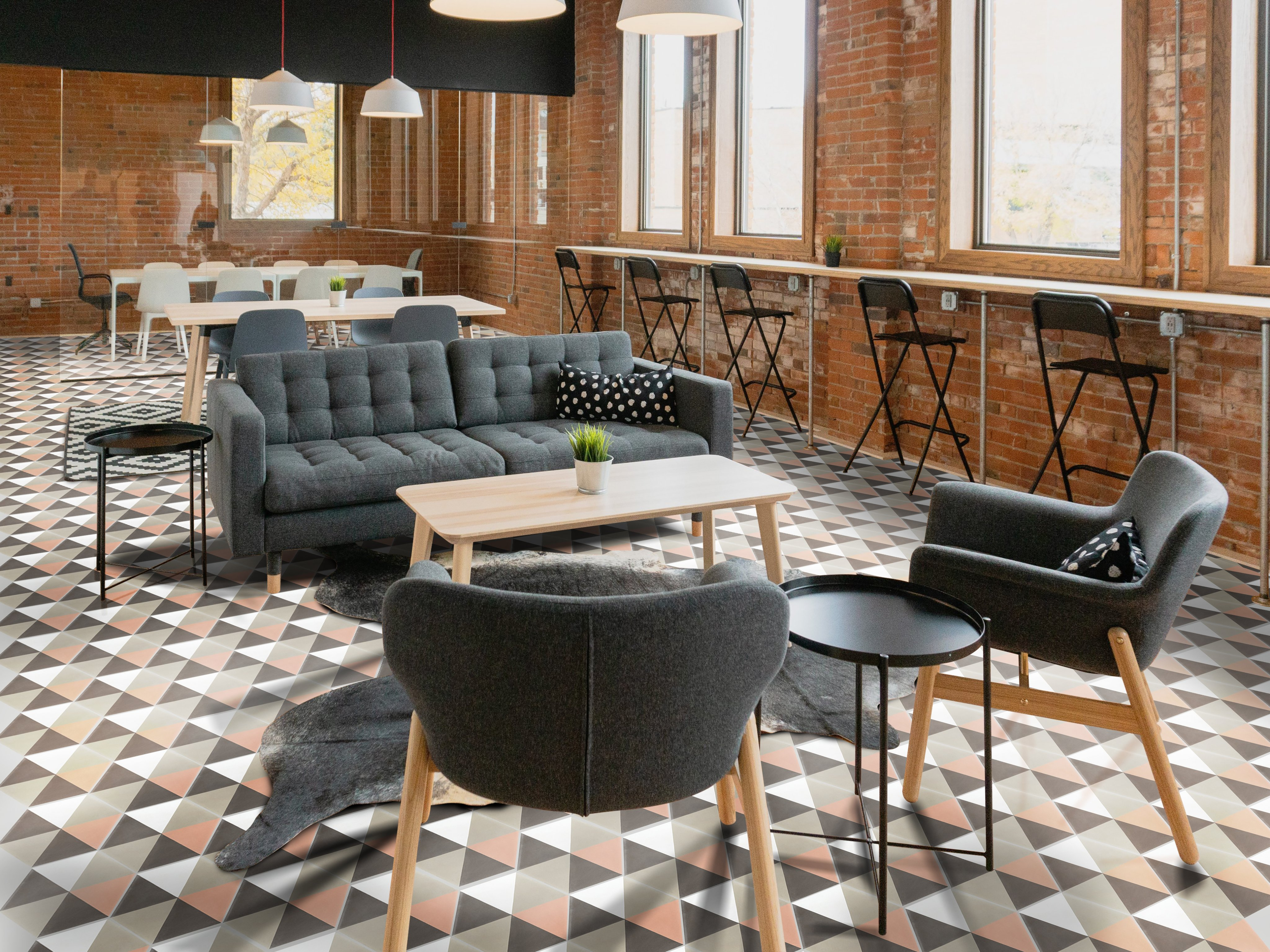 Modern lounge space with gray sofas and chairs, wooden tables, geometric patterned floor, large windows, exposed brick walls, and a row of bar stools along a counter by the windows.