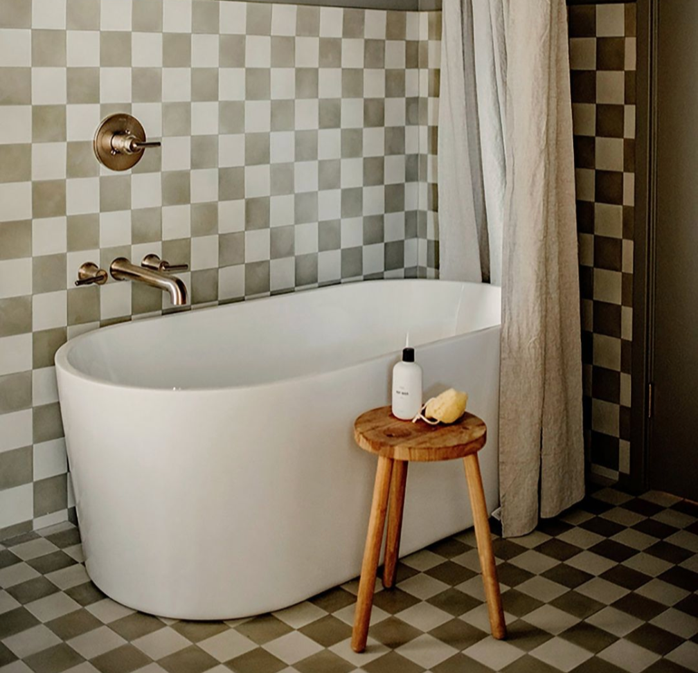 Modern bathroom with a white freestanding tub, checkered beige and white tiled walls and floor, a shower curtain, and a small wooden stool holding a bottle and a sponge.