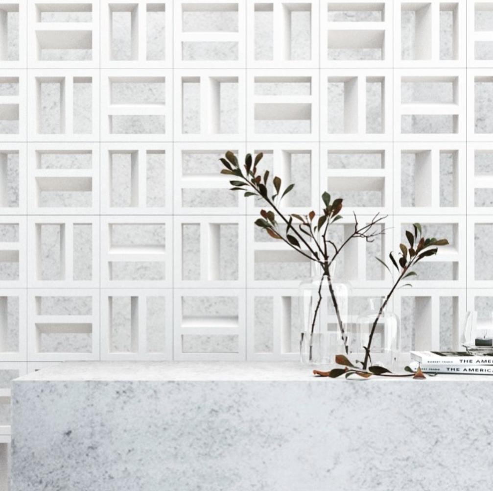 Minimalist scene with a white textured geometric wall, a marble-like countertop, a clear vase holding simple leafy branches, and a few stacked books in the corner. Clean, modern, and airy aesthetic.