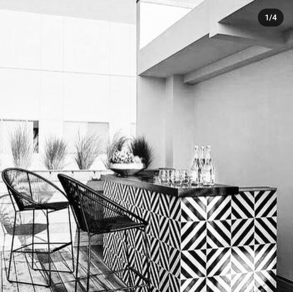 Black and white photo of a modern outdoor bar with geometric-patterned tiles, two wire-frame chairs, and a counter set with bottles and glasses; plants line the background.