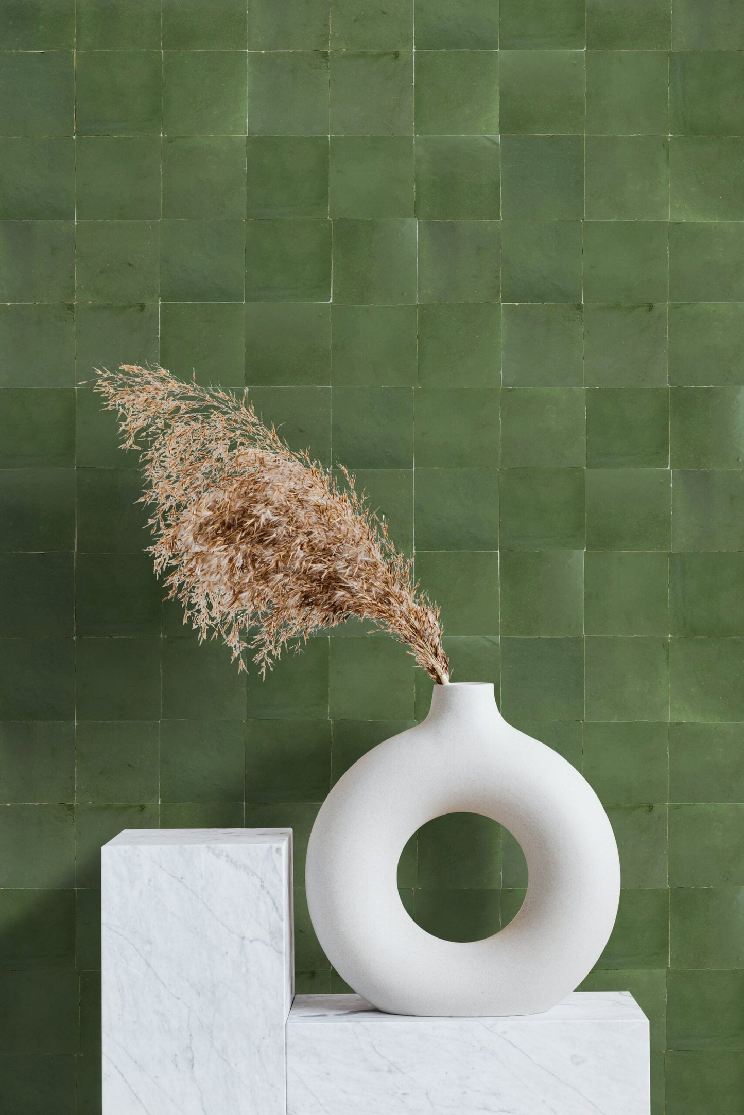 A round white vase with dried grass sits on marble stands before a Moss Green 2X2 tile wall by New York Cement Tile.