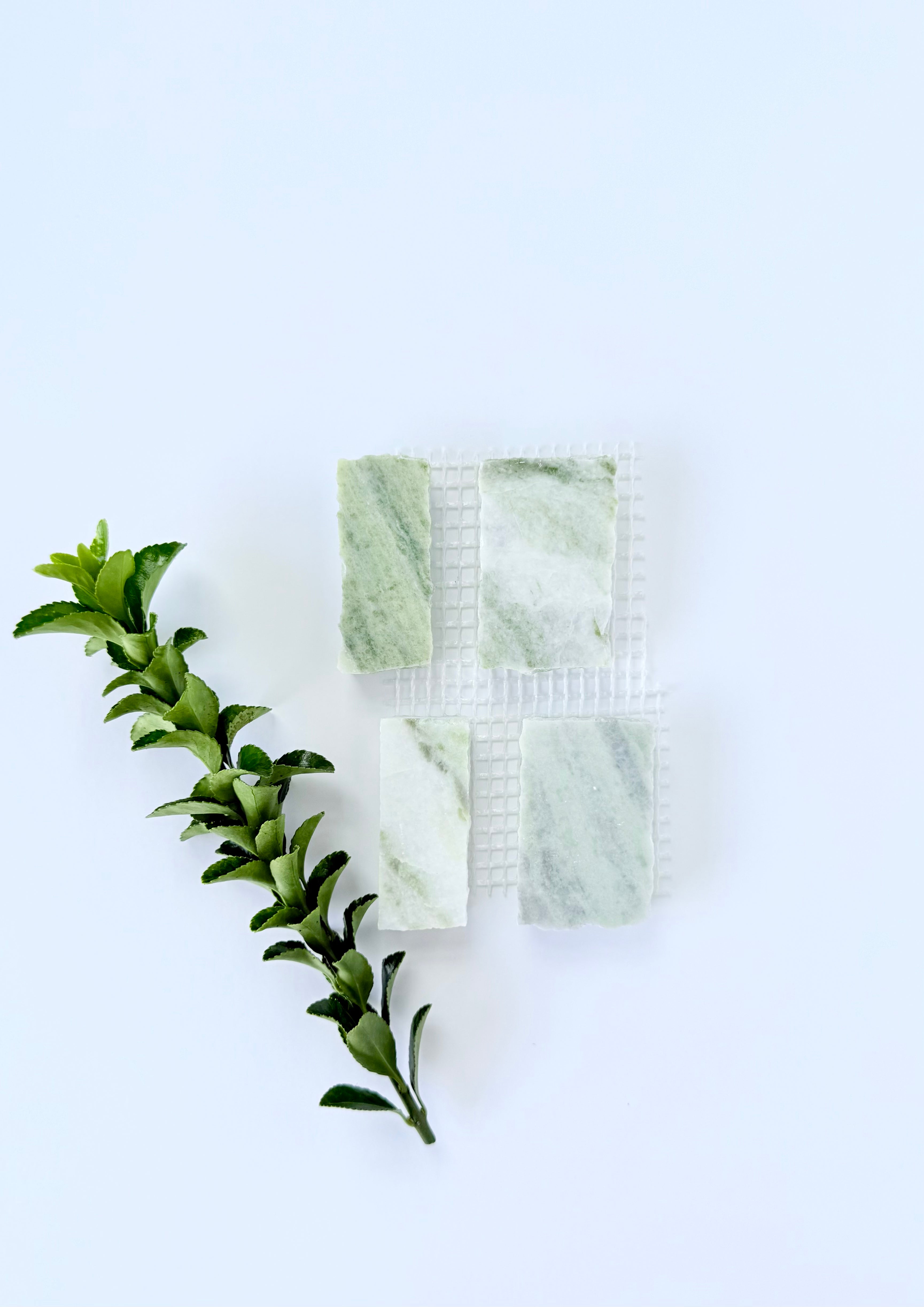 Four Scarpa I Green Vibes marble mesh samples by New York Cement Tile beside green leaves on a white background.