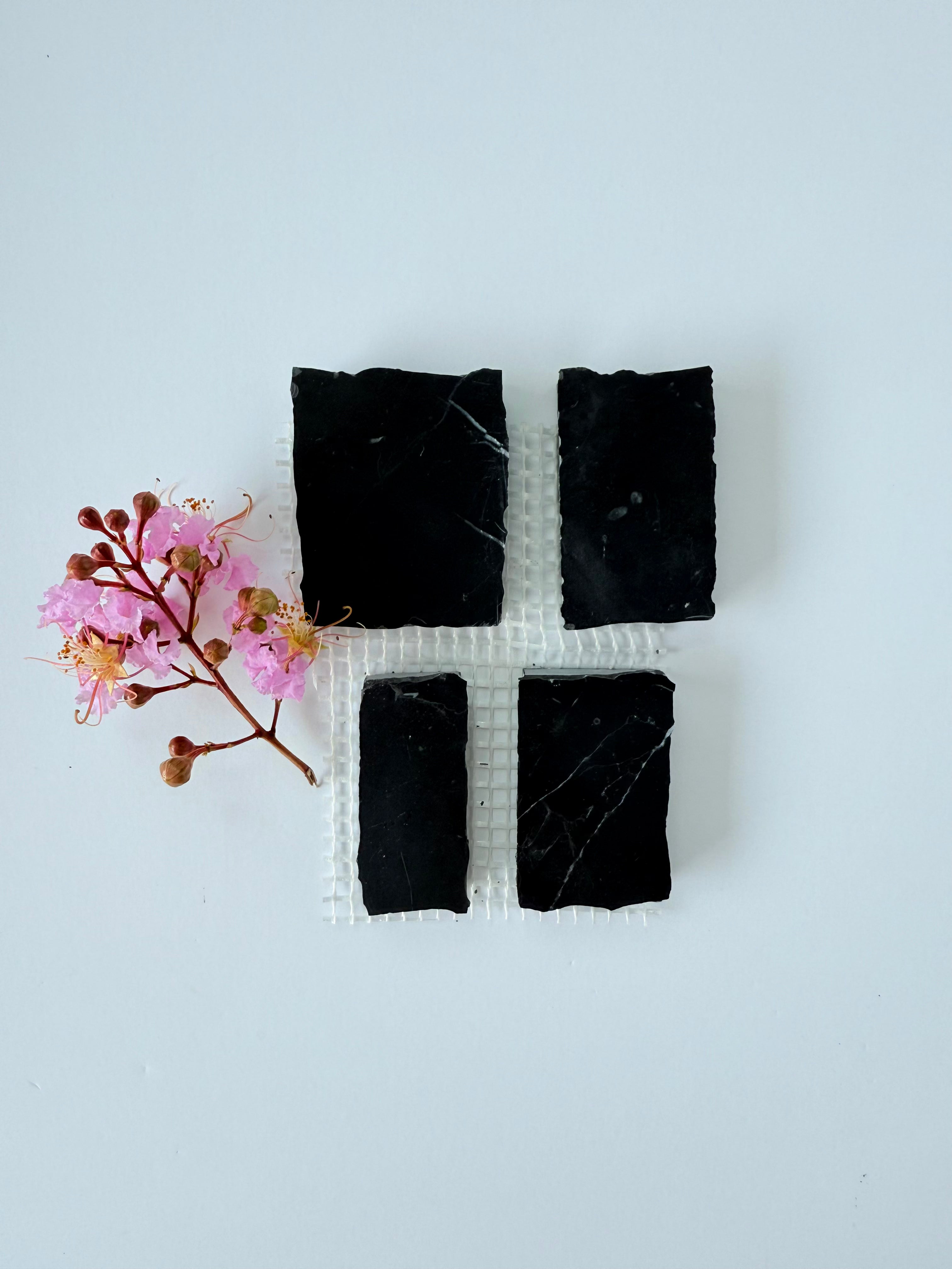 Four Scarpa I Black Mosaic Marble Mesh tiles by New York Cement Tile, with a pink flower sprig on a white background.