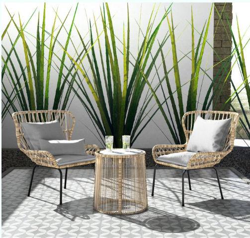 Two wicker chairs and a round table with drinks are set on a tiled patio, backed by tall green plants.