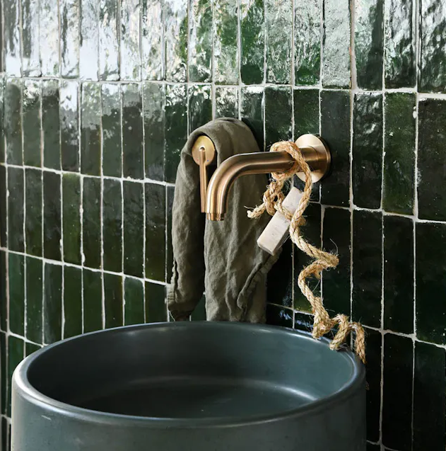 Round dark sink with a brass faucet, soap on a rope, and green tiled wall in the background.