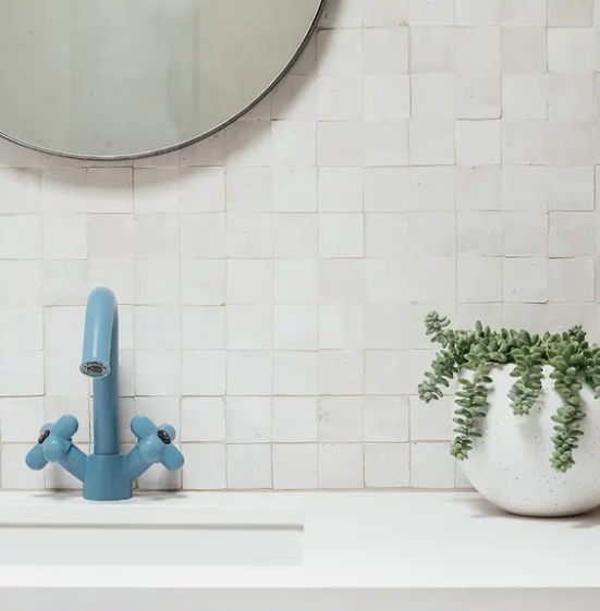 A Frost tile wall by New York Cement Tile features a blue faucet, white sink, round mirror, and potted succulent.