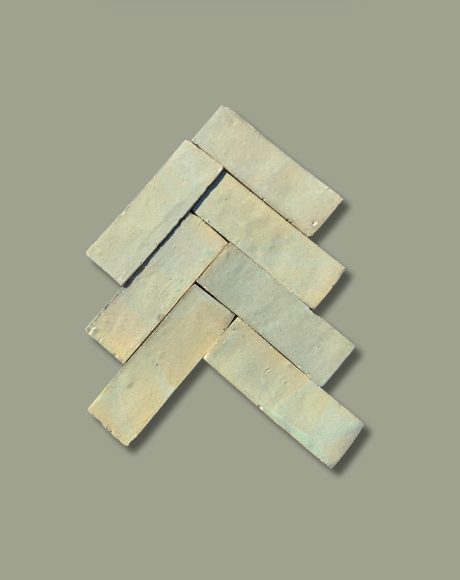 Six Mint Frost (2x6) tiles by New York Cement Tile in a herringbone pattern on a plain, muted green background.