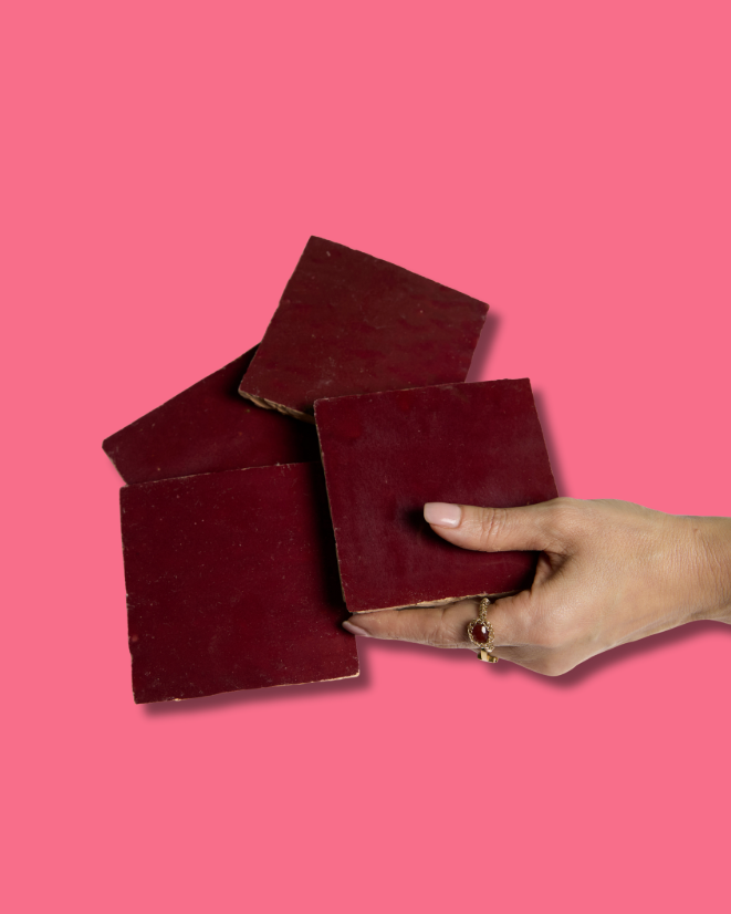 A hand holds a stack of New York Cement Tile's Glazed Sangria tiles in dark red against a pink background.