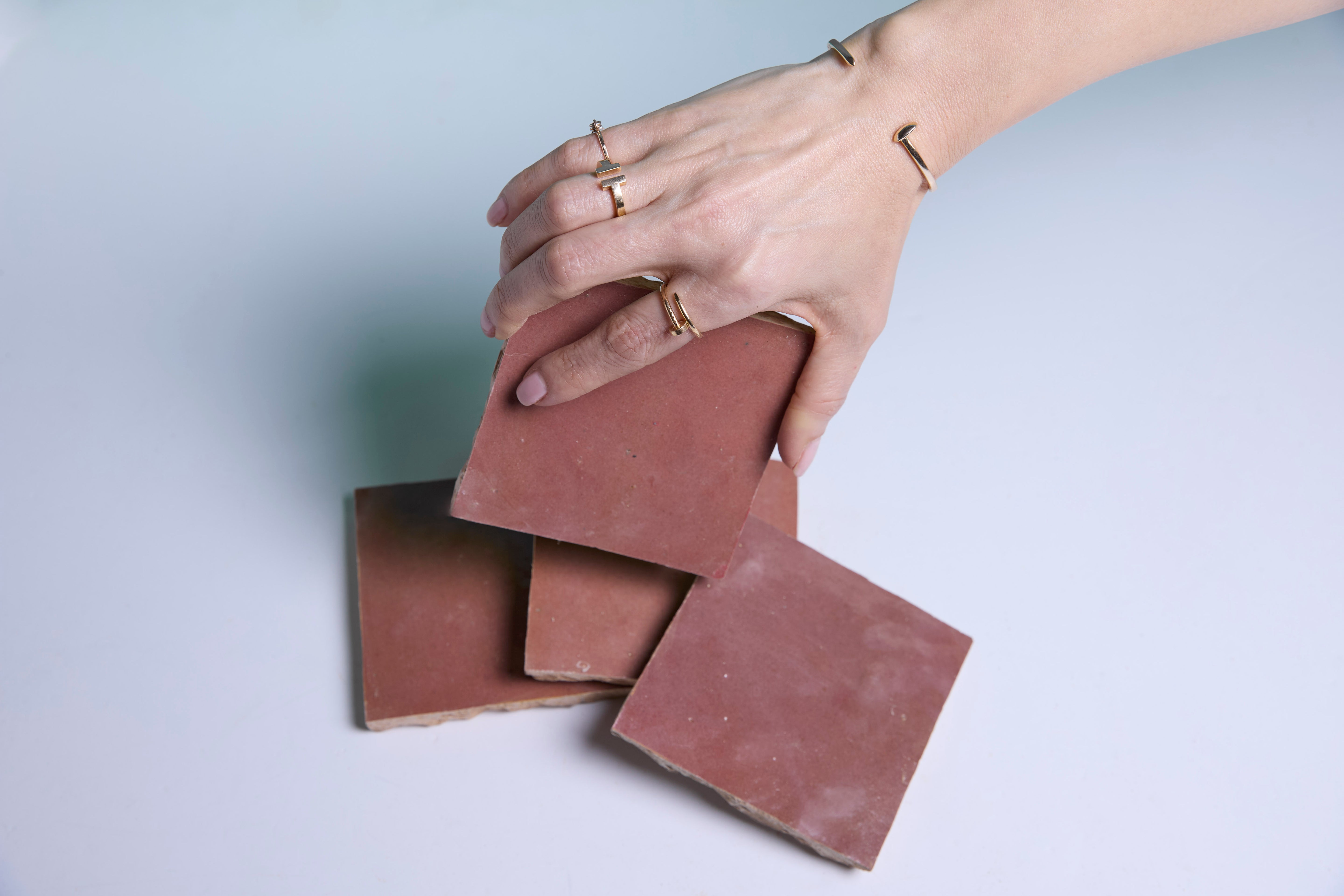 A hand wearing gold rings and bracelets holds a reddish-brown tile above three similar tiles on a white surface.