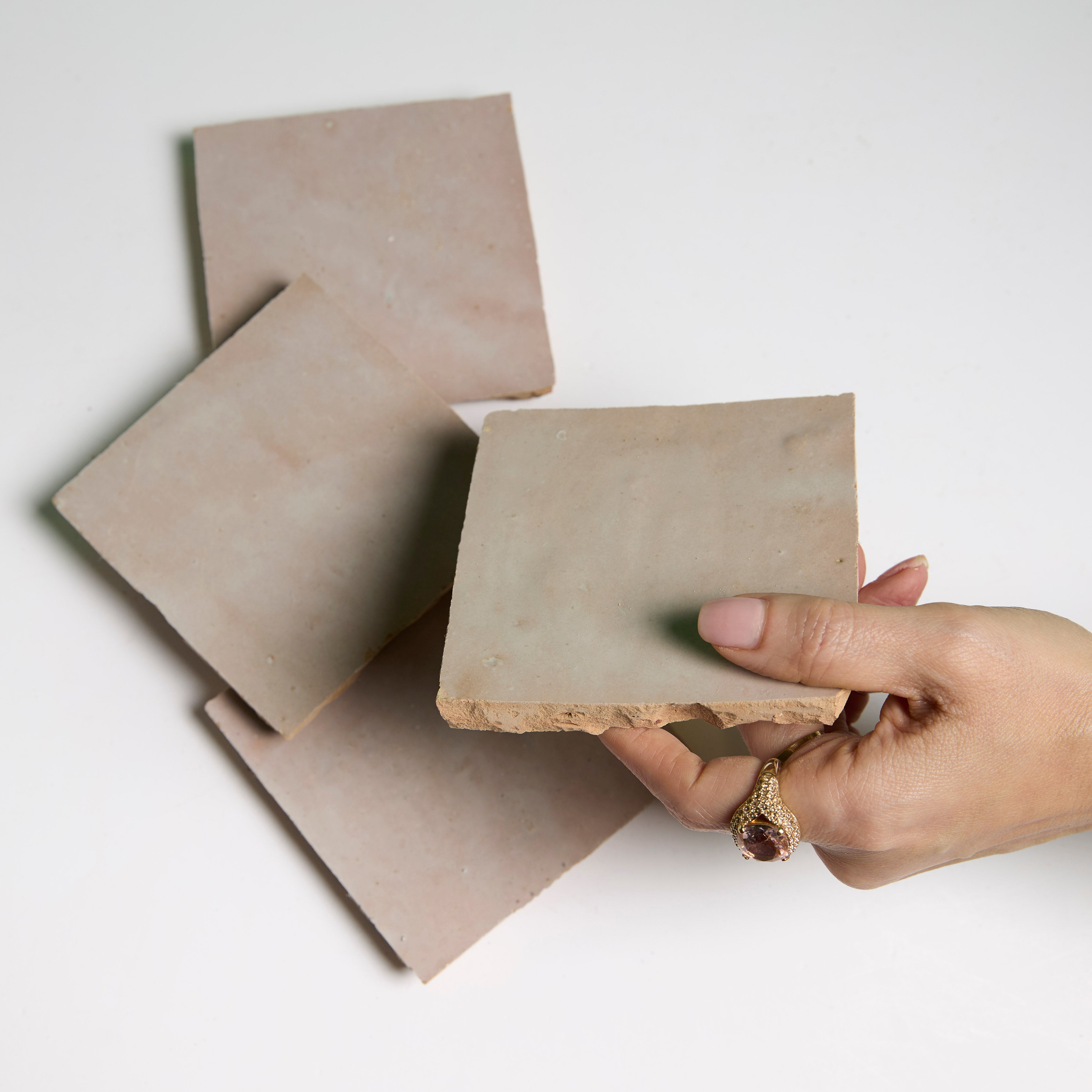 A hand with a gold ring holds a New York Cement Tile Blushed Glazed square tile above three tiles on a white surface.