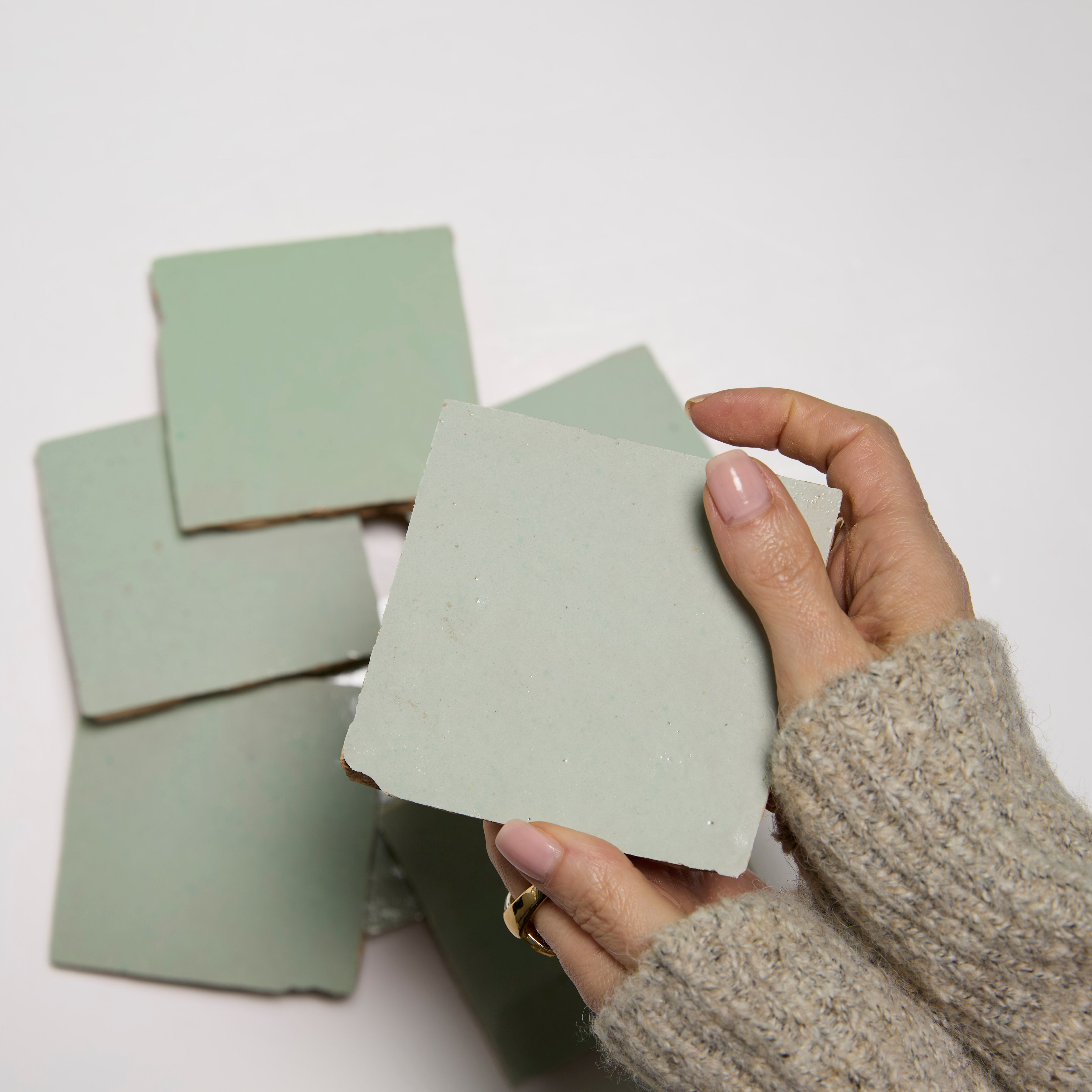 A hand holds a Mint Frost tile by New York Cement Tile above a stack of handcrafted tiles on a white surface.