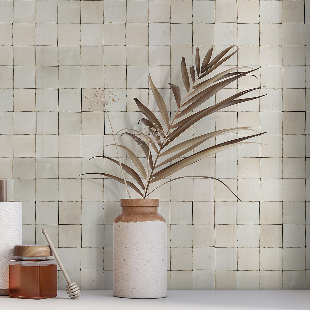 A stone vase with dried palm leaves sits beside a honey jar, against New York Cement Tile’s Pearl Grey 2X2 backsplash.