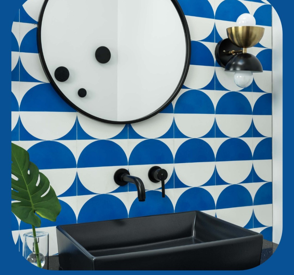 Modern bathroom with blue and white geometric tiles, round mirror, black sink, black faucet, and a wall sconce.
