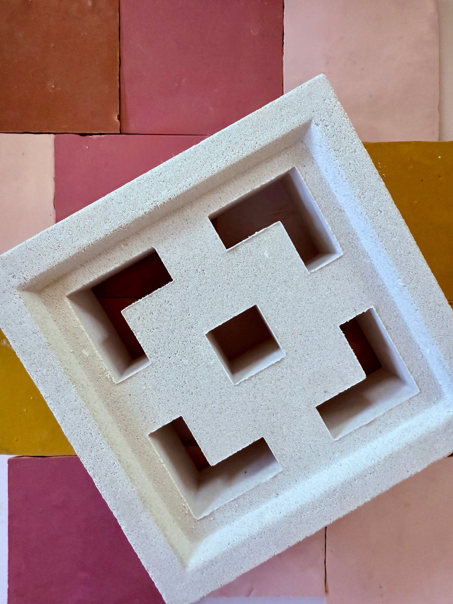 A white, geometric breeze block with square and cross-shaped cutouts is placed diagonally on a background of pink, red, and yellow square tiles.