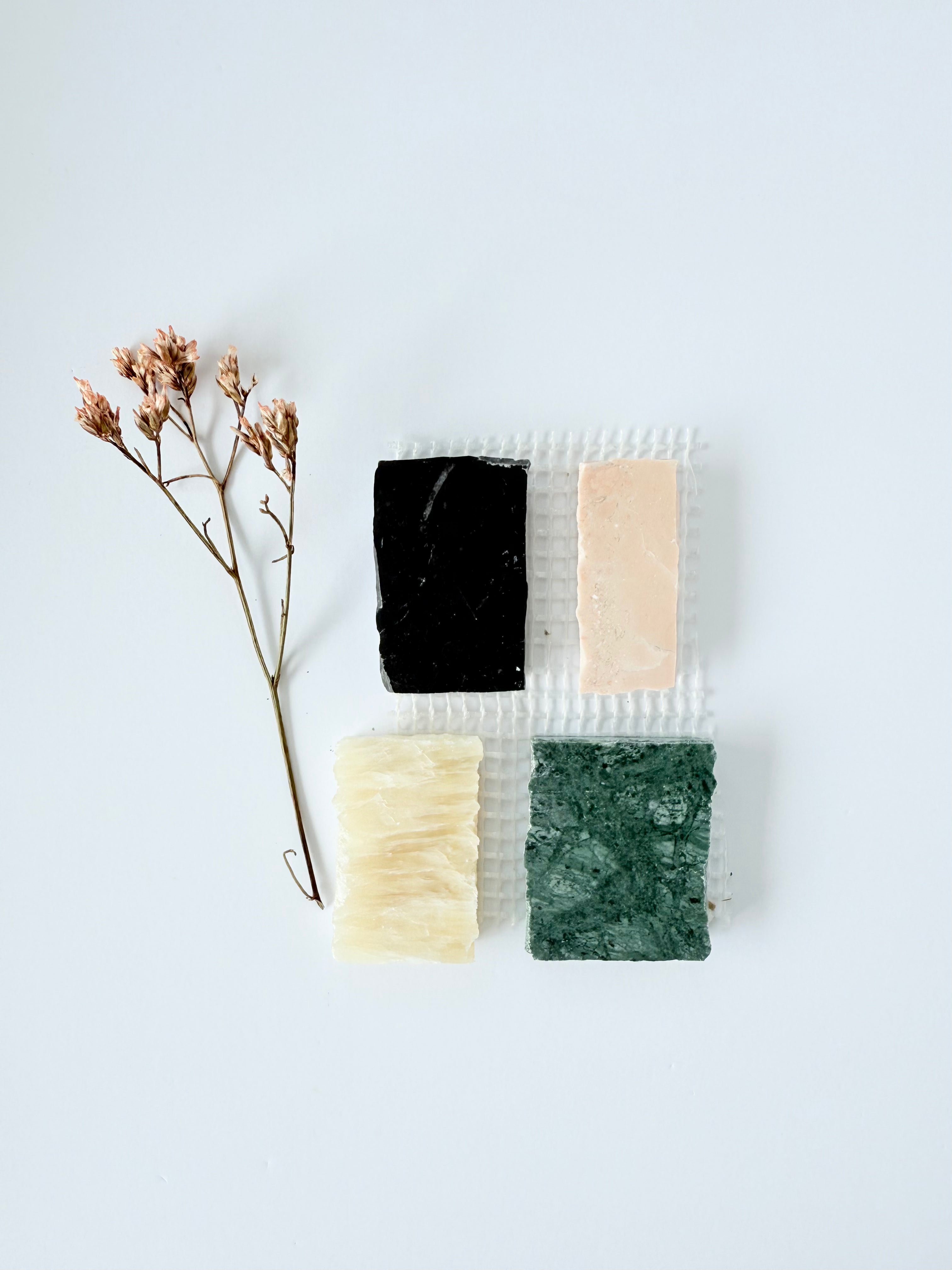 Four rectangular soap bars in black, pink, cream, and green are arranged on a textured white surface with a dried sprig of flowers beside them, all set against a plain white background.