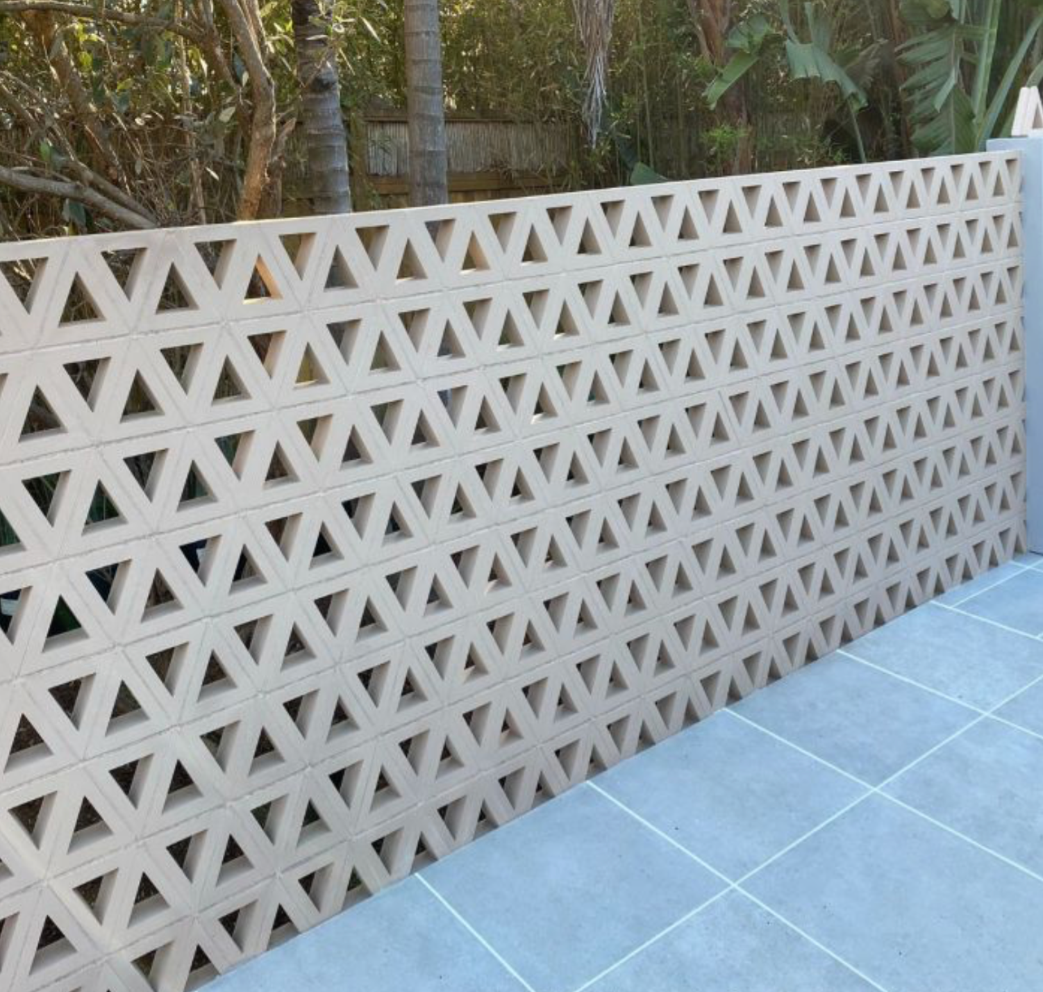A decorative wall made of beige concrete blocks with repeating triangle cutouts stands beside a tiled patio, with trees and plants visible in the background.