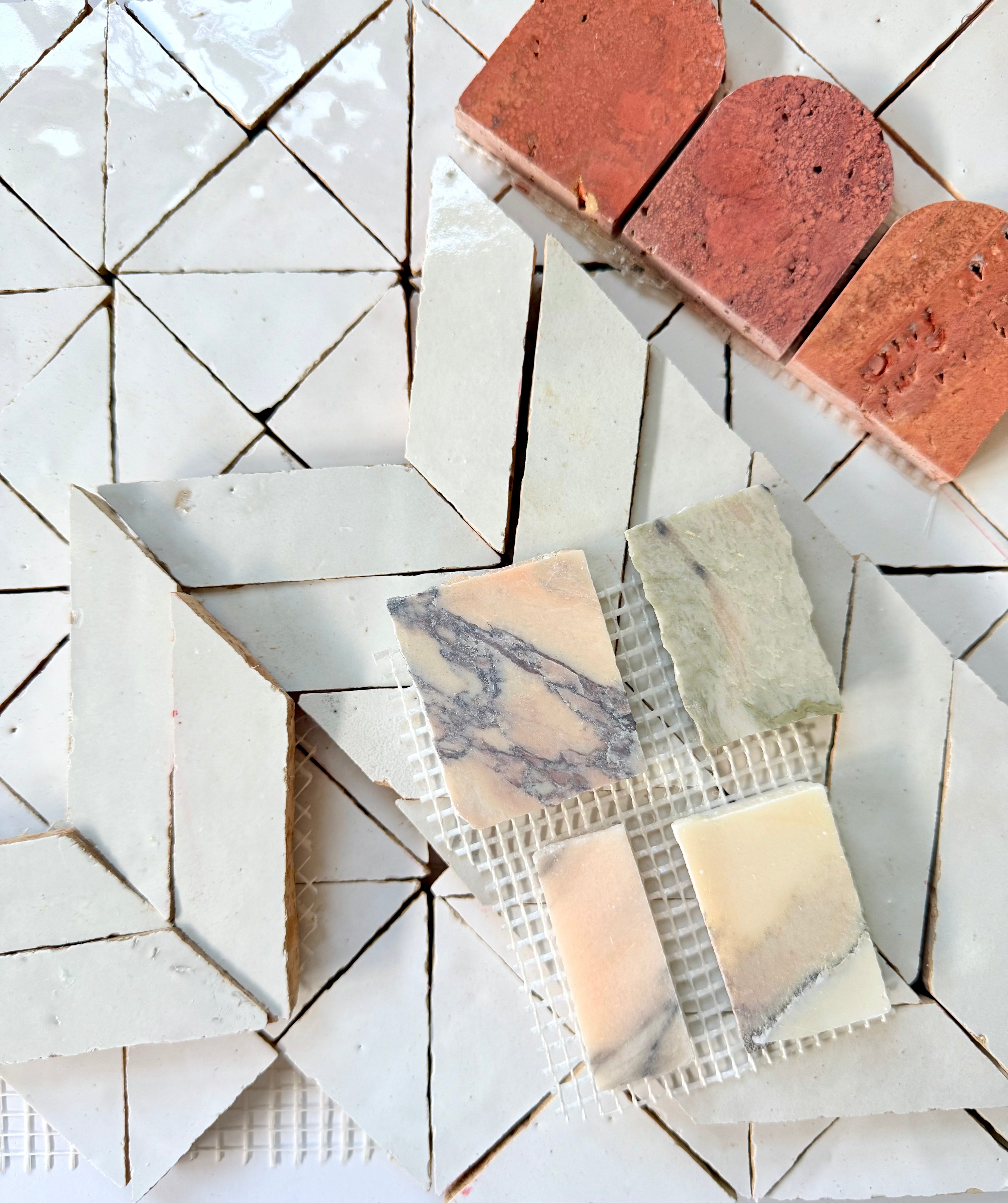 Assorted tiles in various shapes, colors, and patterns are arranged on a geometric white tiled surface, including red bricks, cream chevrons, and marble samples on mesh.