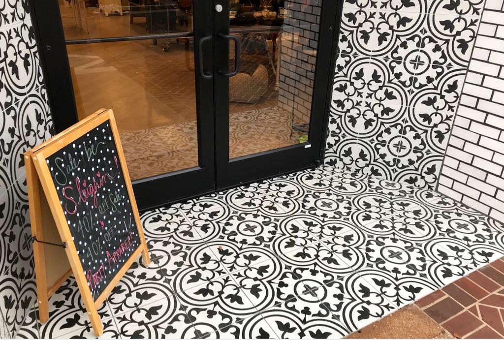 Black double doors with patterned black-and-white tile and a chalkboard sale sign outside a store entrance.