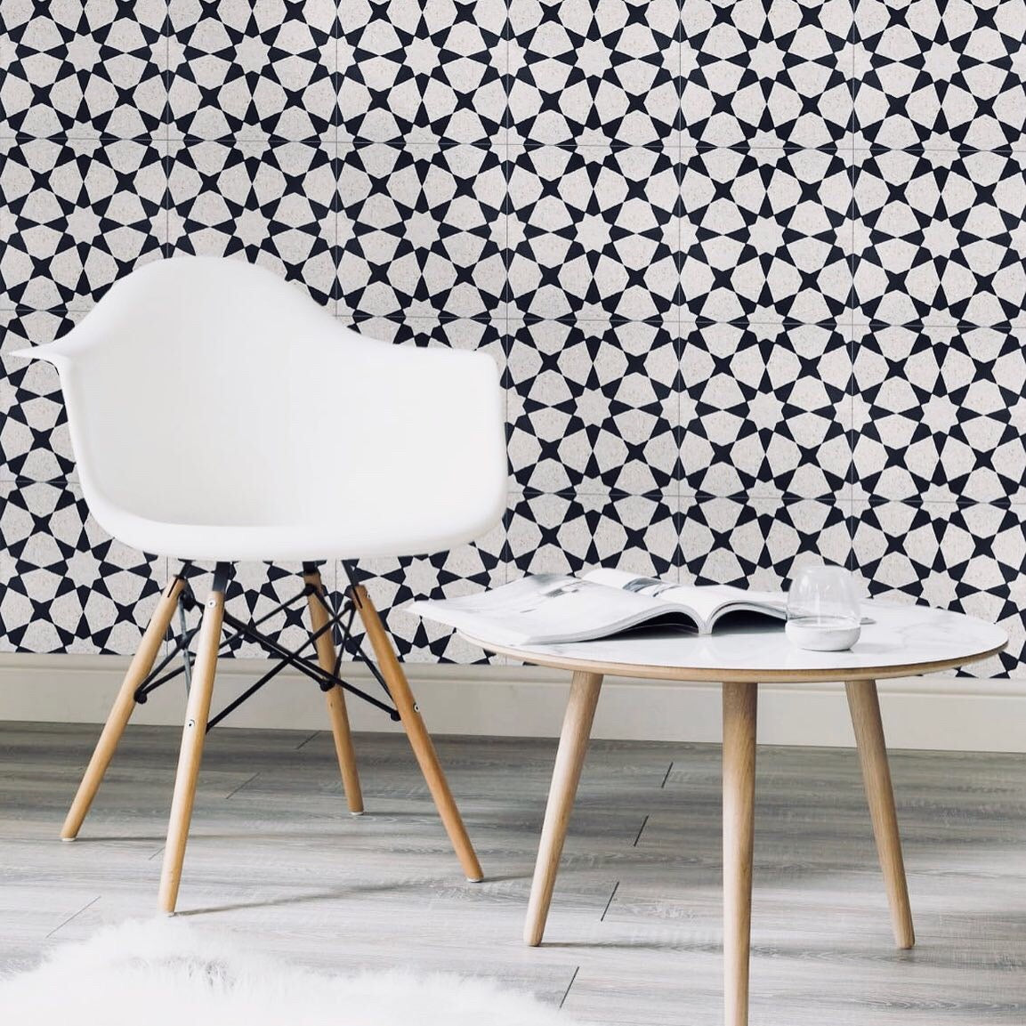 A white chair and round table with an open book in front of a black and white geometric patterned wall.