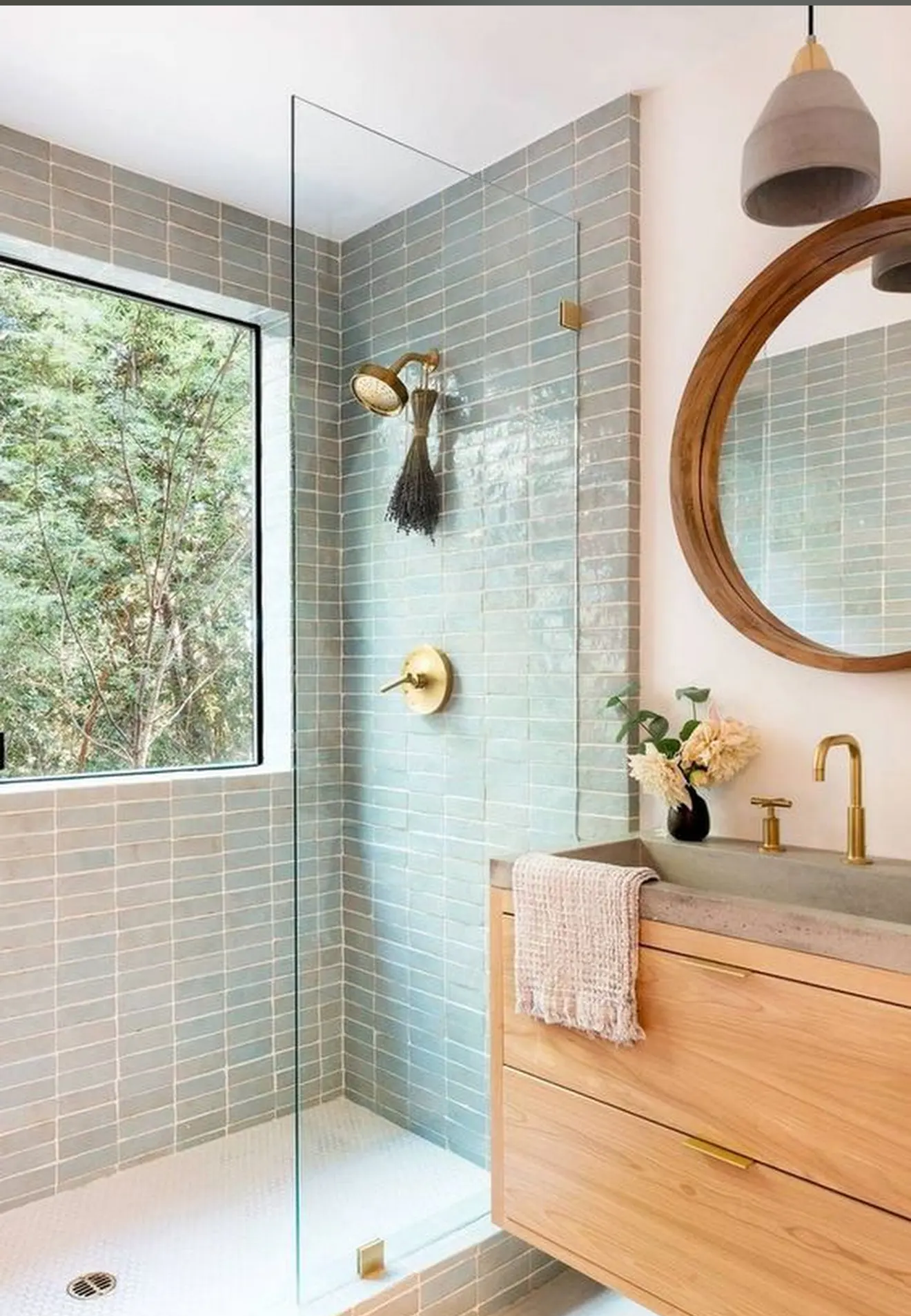 Modern bathroom with glass shower, light wood vanity, round mirror, gold fixtures, and a large window with a view of trees.