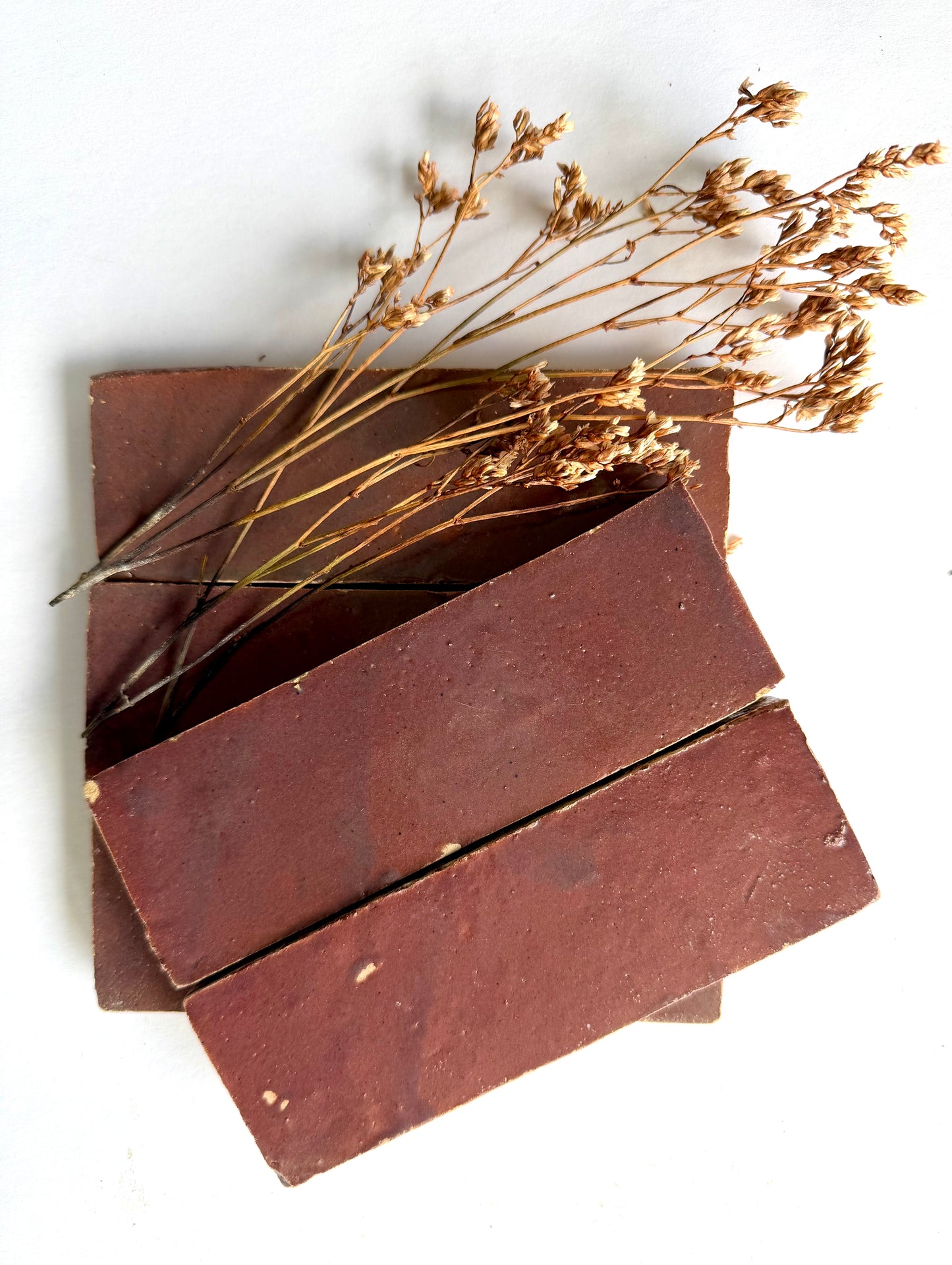 Three Thistle (2x6) brown subway tiles by New York Cement Tile stacked with dried grass atop a white background.