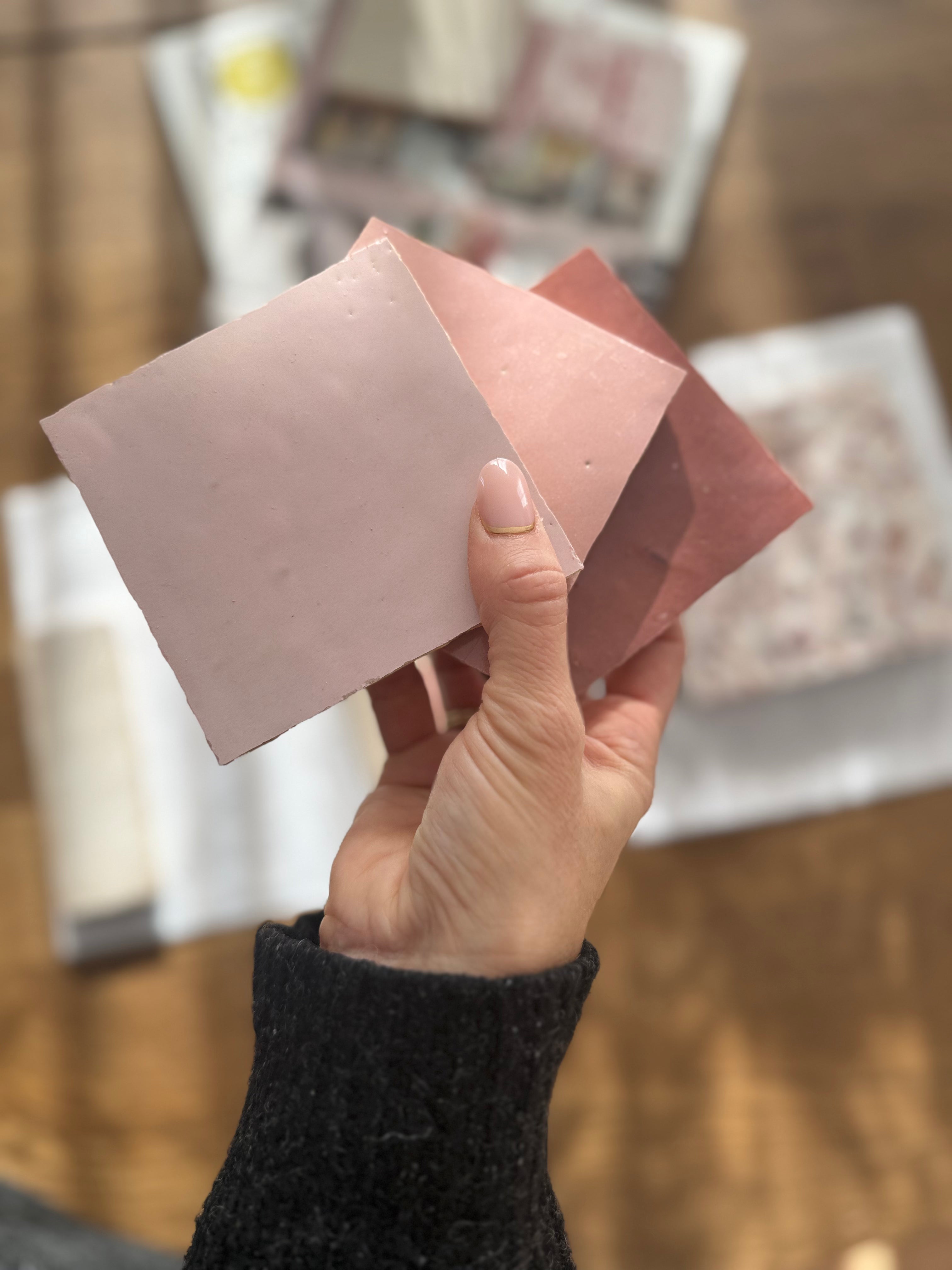 A hand holds three Vintage Rose tiles by New York Cement Tile above a table with design materials in soft natural light.