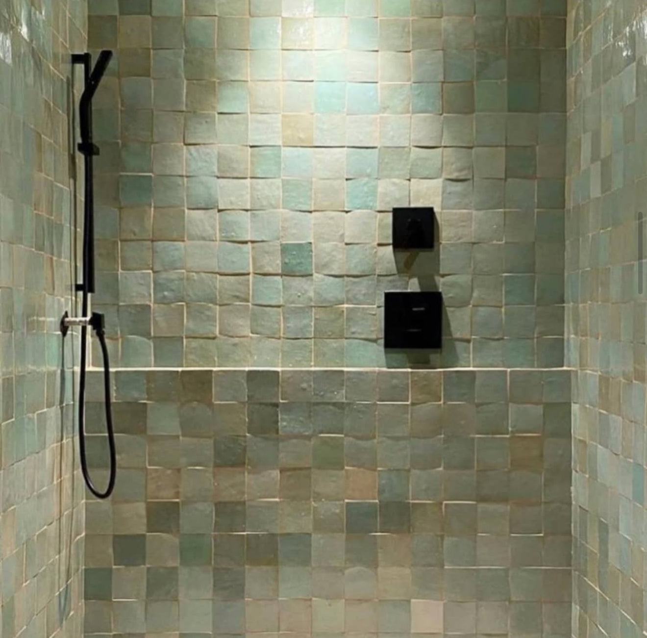 Shower with greenish square tiles, black handheld showerhead, and two black wall controls.