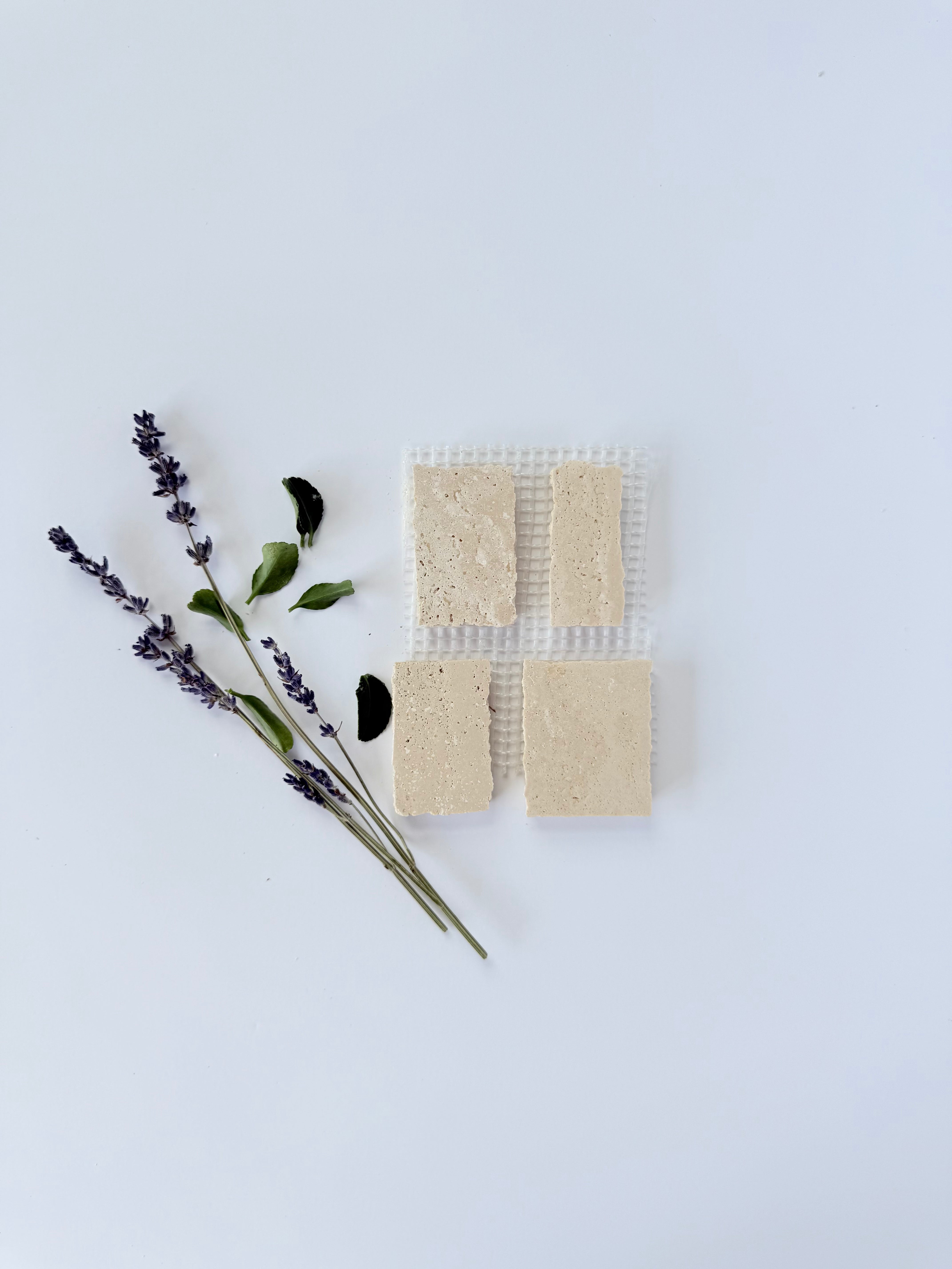 Four rectangular beige Scarpa I Travertine Mosaic Marble Mesh tiles by New York Cement Tile, on white mesh with leaves.