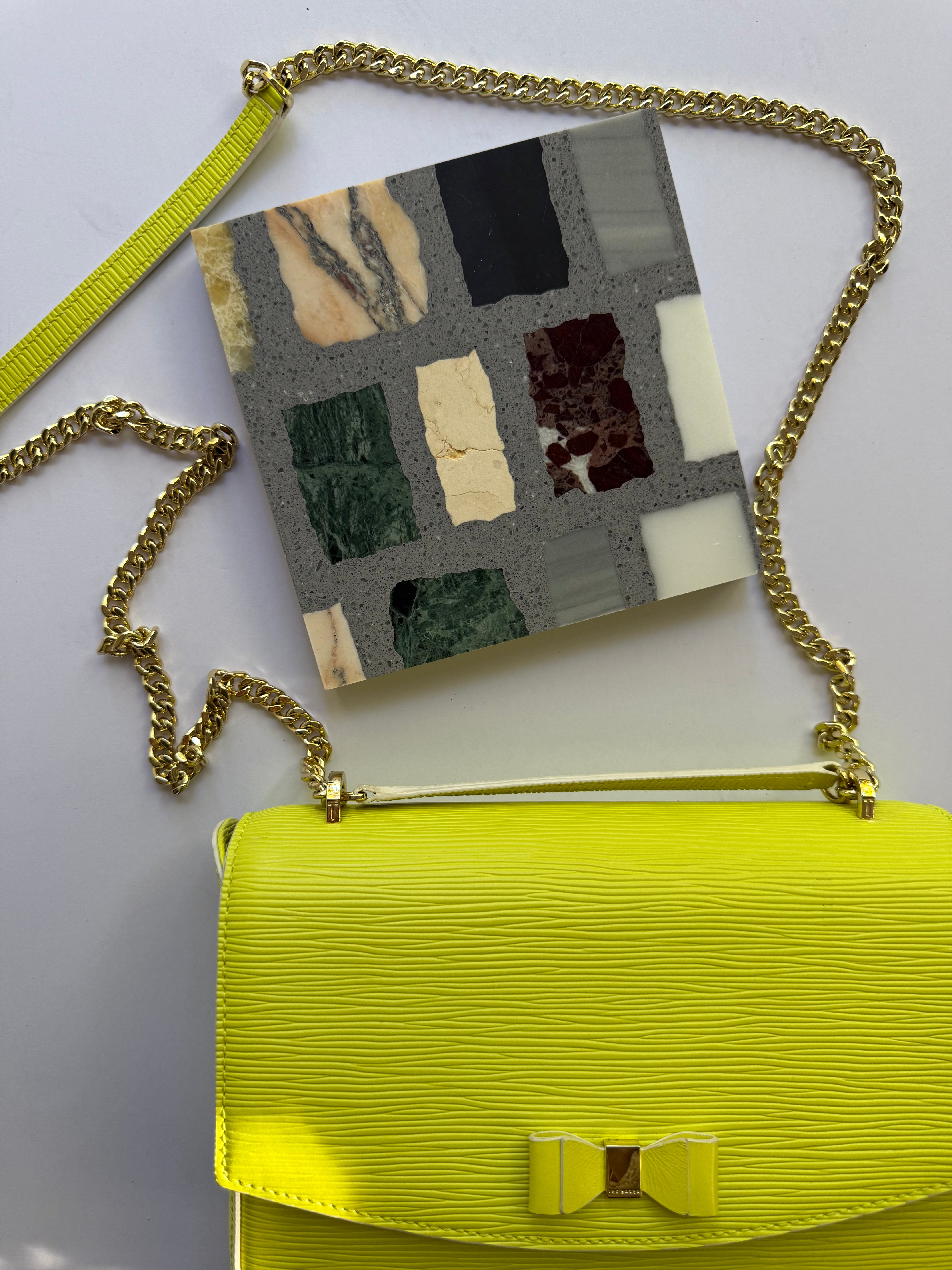 Yellow purse with gold chain strap next to New York Cement Tile's Carlo Scarpa- Multicolor Dark tile sample on white background.
