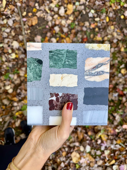 A hand with red nails holds a Carlo Scarpa- Multicolor Dark tile by New York Cement Tile over fallen leaves.