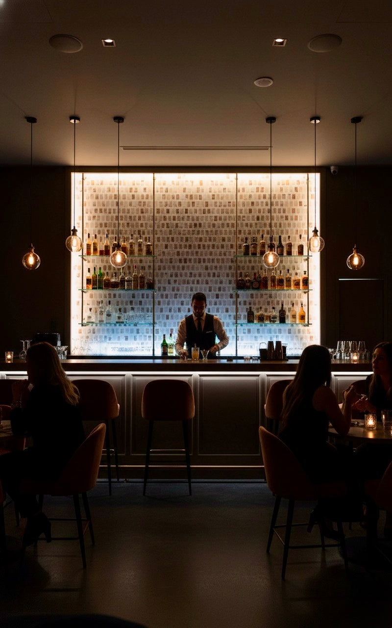 A bartender stands behind a bar with Carlo Scarpa - Mountain High tiles by New York Cement Tile in a modern, dim lounge.