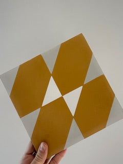 A hand holds the NL20-4D tile by New York Cement Tile, showcasing mustard yellow, gray, and white geometric patterns.