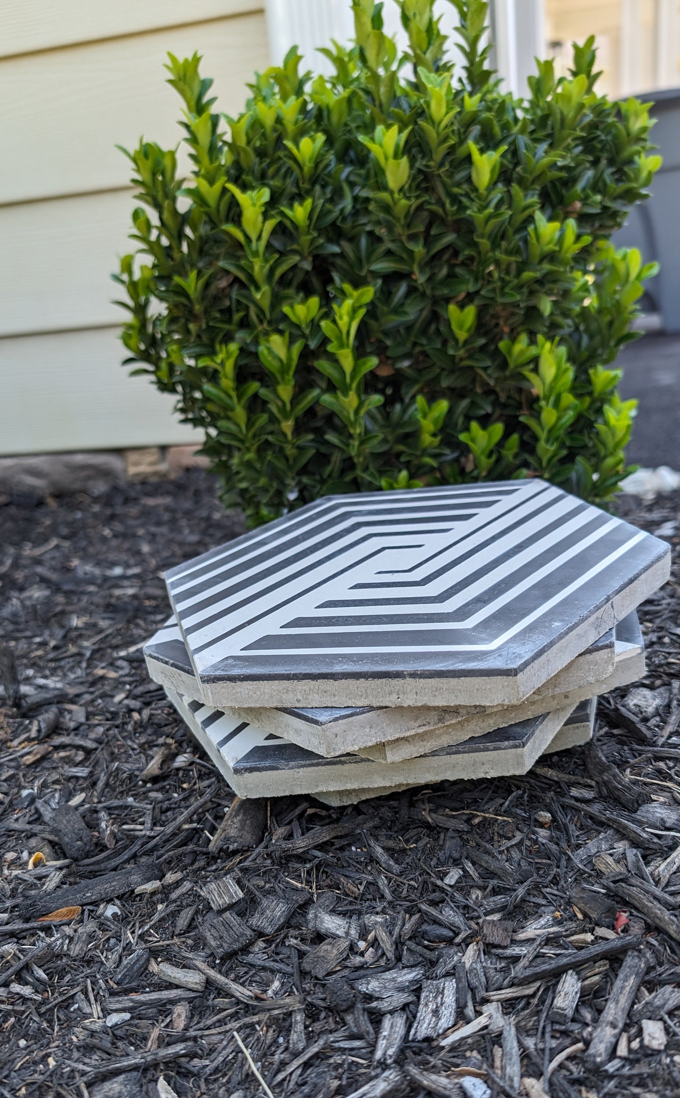 Three NL23-7 hexagon tiles by New York Cement Tile with black and white geometric patterns stacked on mulch by a shrub.