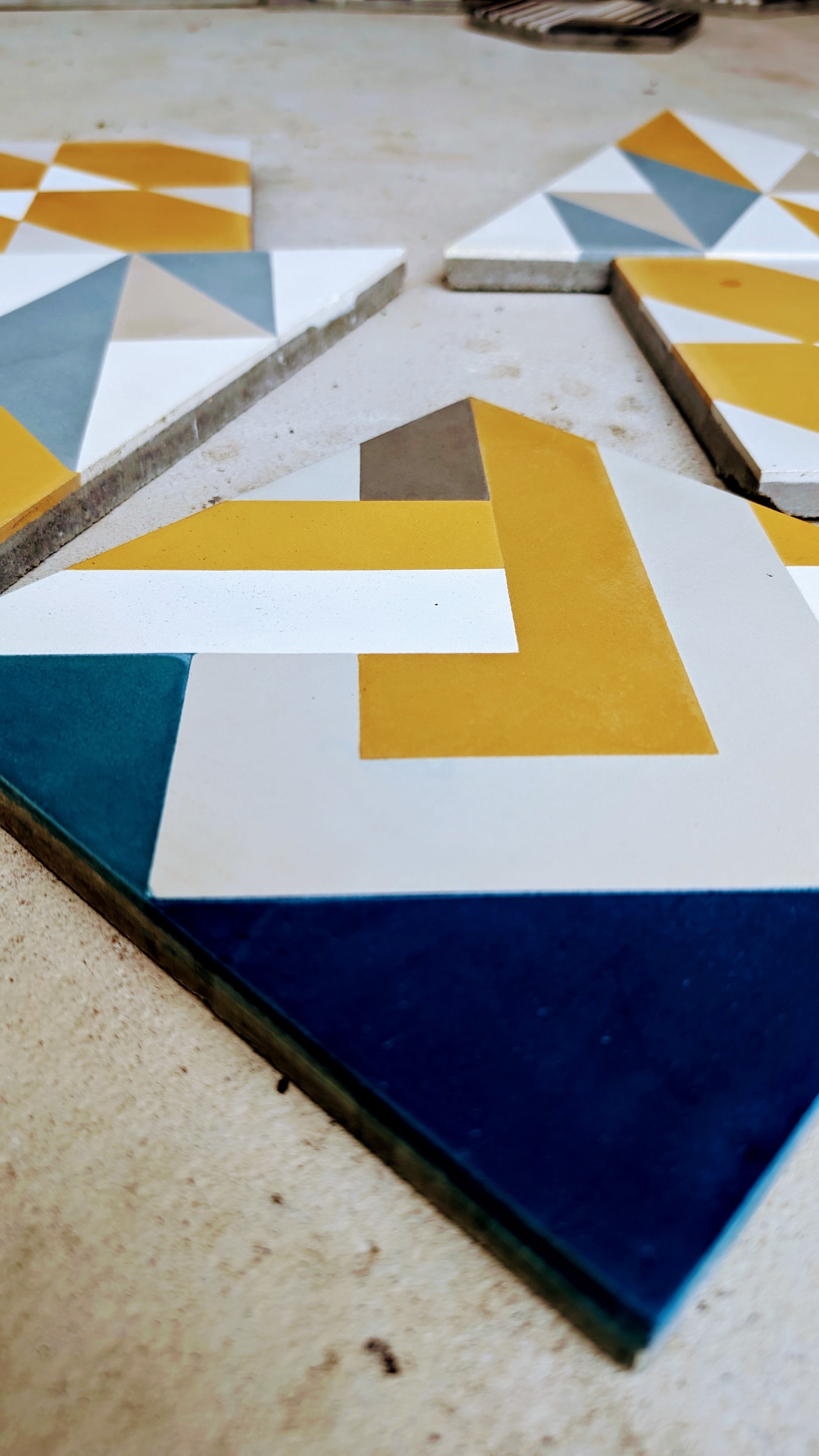 NL20-1A geometric tiles by New York Cement Tile in yellow, blue, white, and gray are set on a light-colored surface.