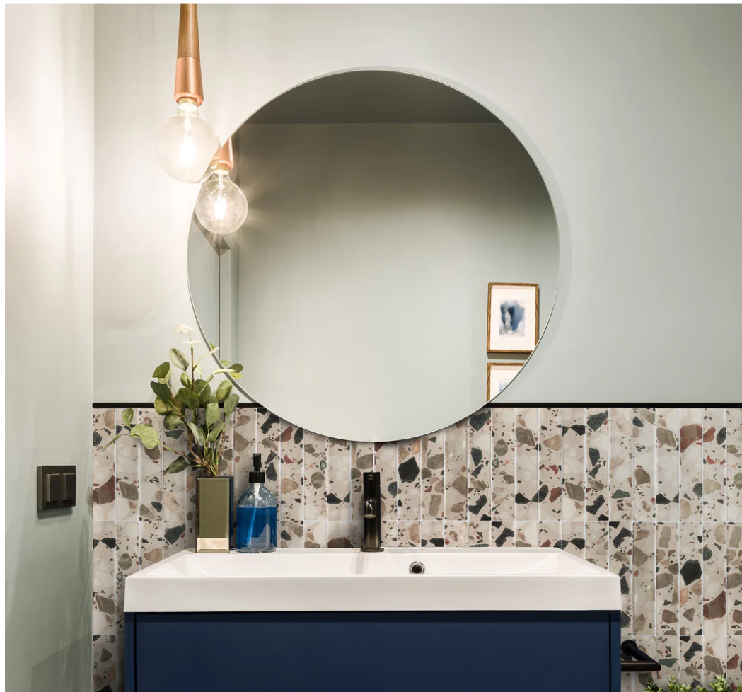 Modern bathroom with round mirror, terrazzo tiles, blue vanity, pendant lights, and a small plant on the sink.