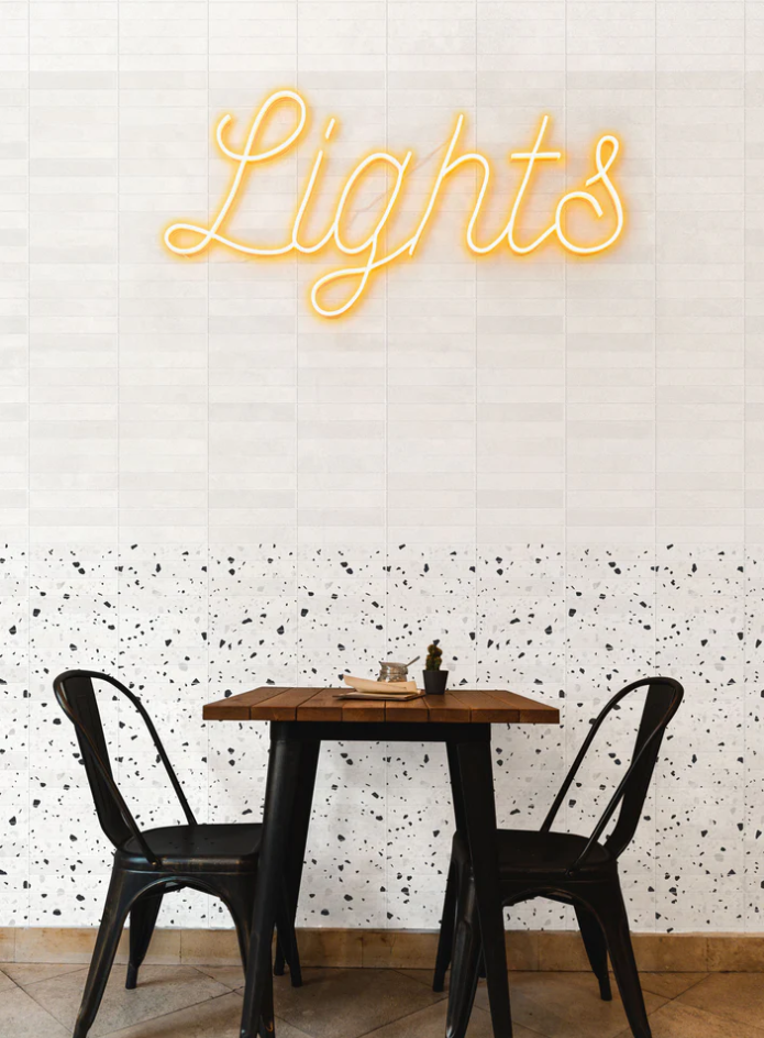 Small table with two black chairs under yellow neon lights on New York Cement Tile’s CORRAZZO-NEGRO ceramic tile wall.