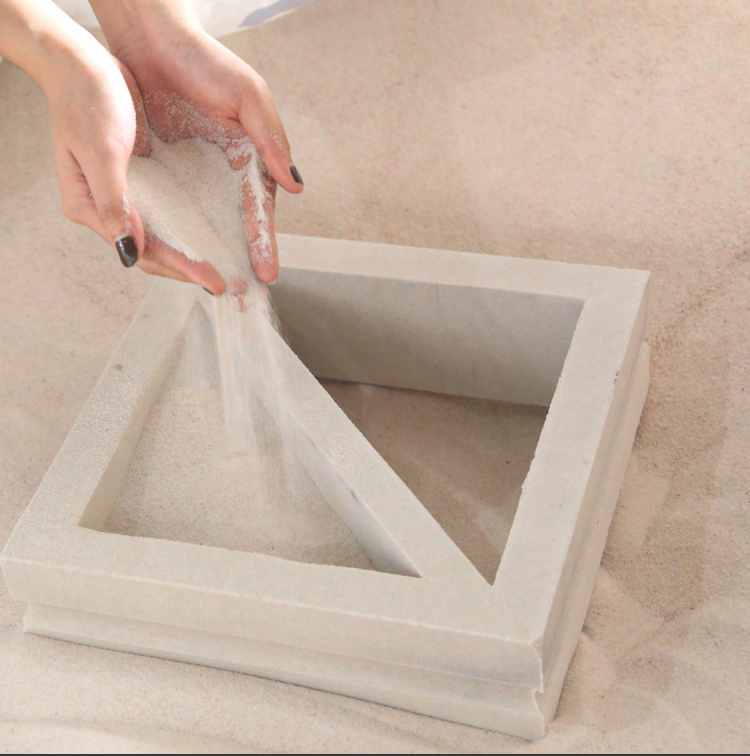Hands pouring sand into a square frame with a diagonal divider, all set on a sandy surface.