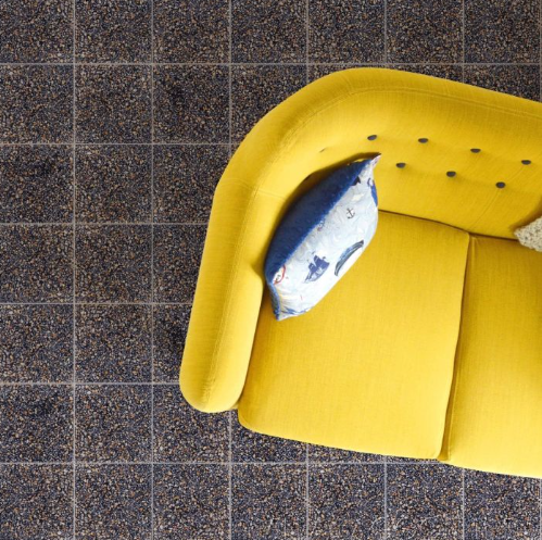 Yellow armchair with blue button details and a cushion, on a speckled tiled floor, viewed from above.