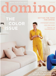 A woman in a yellow jumpsuit stands by a beige sofa with pillows in a bright, modern living room; domino magazine cover.