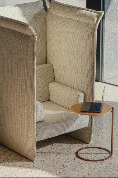 A cozy beige enclosed chair with a small round side table holding an open laptop, next to a sunlit window.