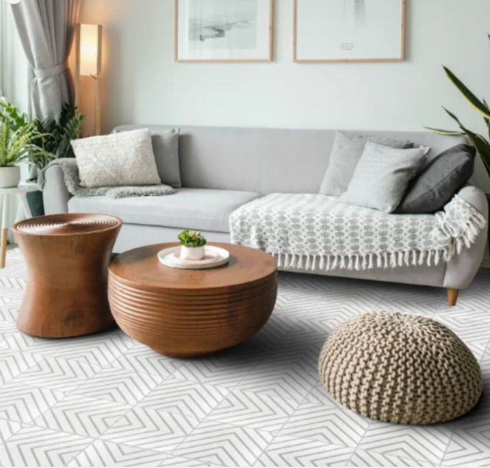 Modern living room with a gray sofa, round wooden tables, knit pouf, plants, and patterned white floor.