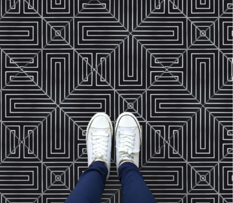 Person wearing white sneakers standing on a black and white geometric patterned floor with intersecting straight lines.