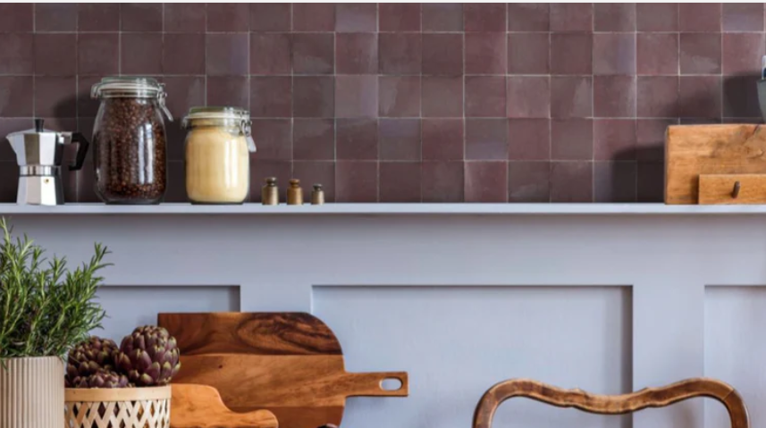 A kitchen shelf with jars, a coffee maker, wooden boards, artichokes, and a tiled backsplash in earthy tones.