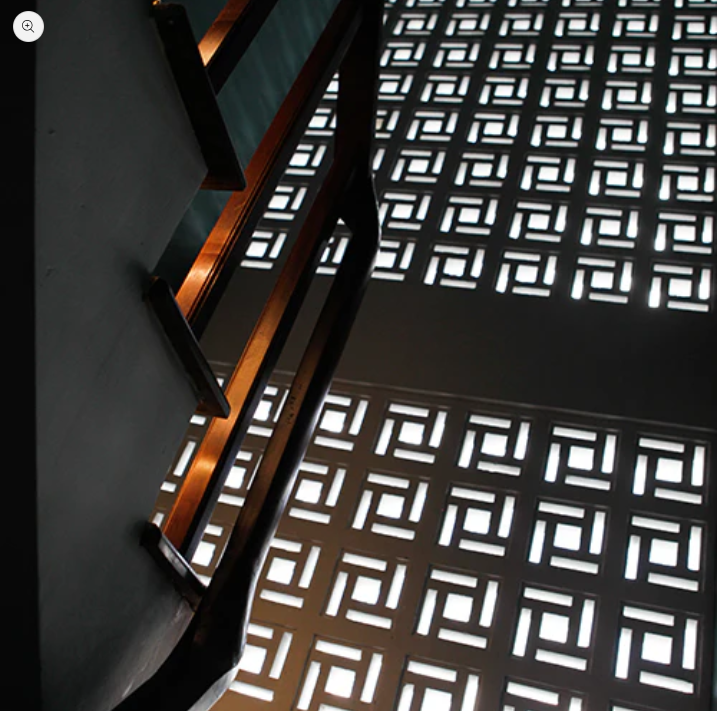 Sunlight filters through geometric-patterned windows, casting light squares on a wall beside a stairway with a wooden railing.