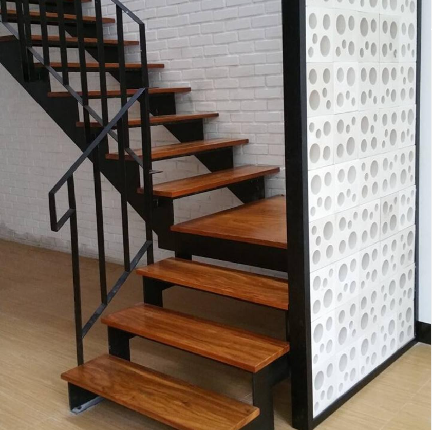 Modern wooden staircase with black metal railing next to a white perforated partition and brick wall background.