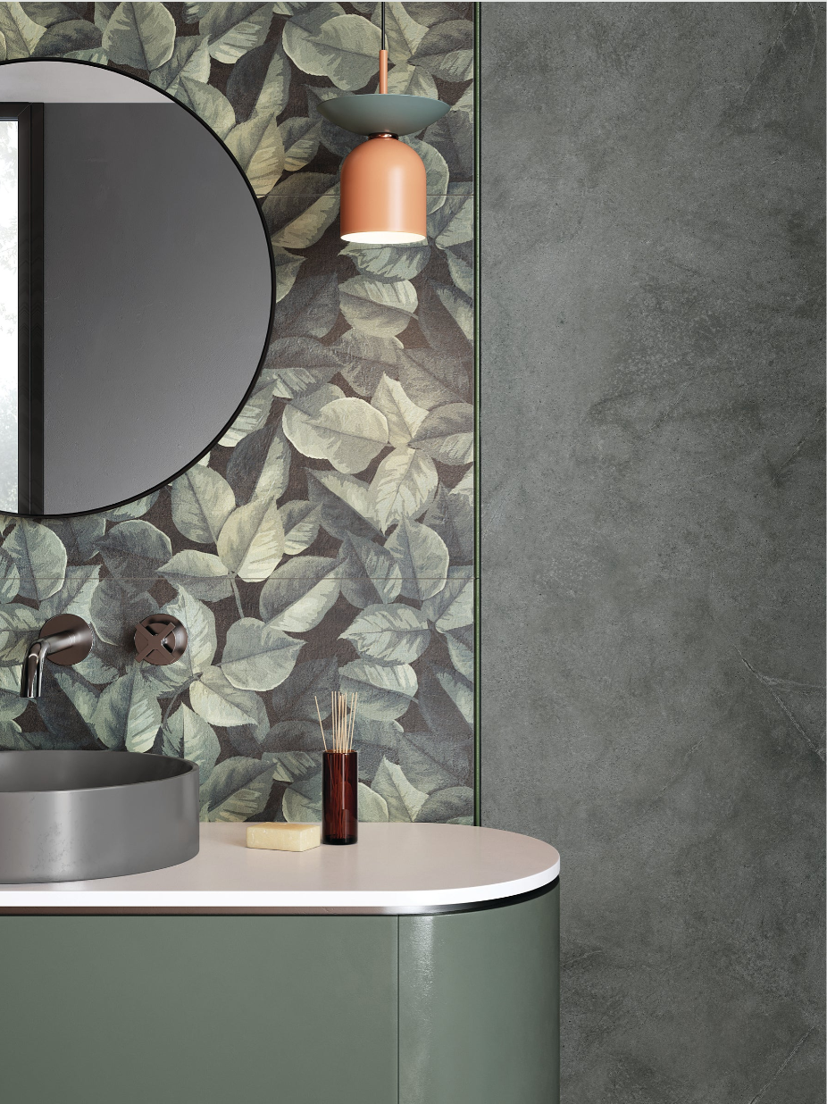 Modern bathroom with leafy wallpaper, round mirror, gray sink, orange pendant light, and green cabinets.