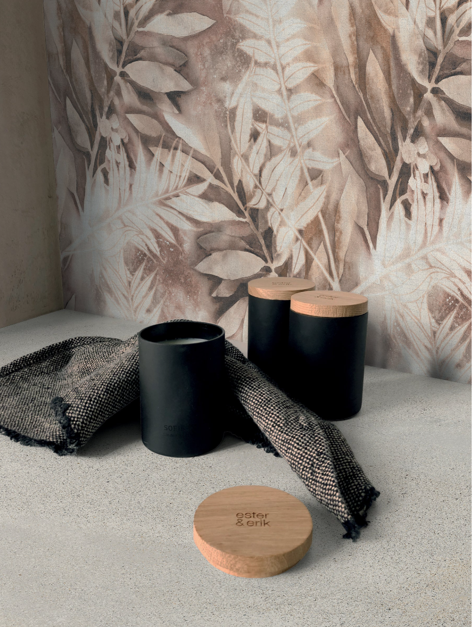 Black jars with wooden lids and a textured cloth on a light surface, against a brown leafy patterned wall.